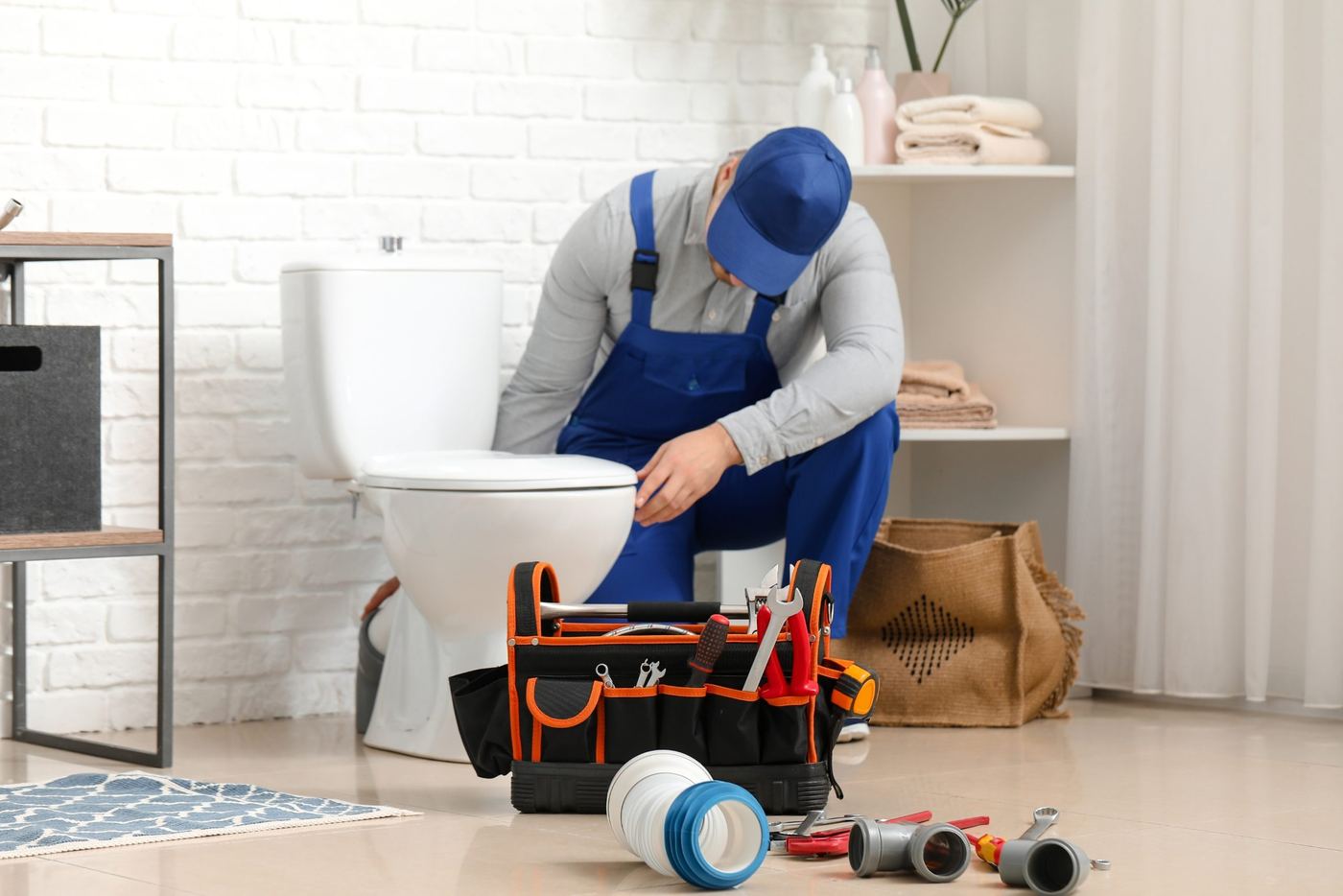 A plumber in blue overalls working on a toilet, with a tool bag and fittings on the floor